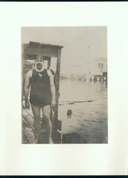 Nasser Mann in Badeanzug steht auf einem Holzsteg neben einem kleinen See mit einem schwimmenden Mann